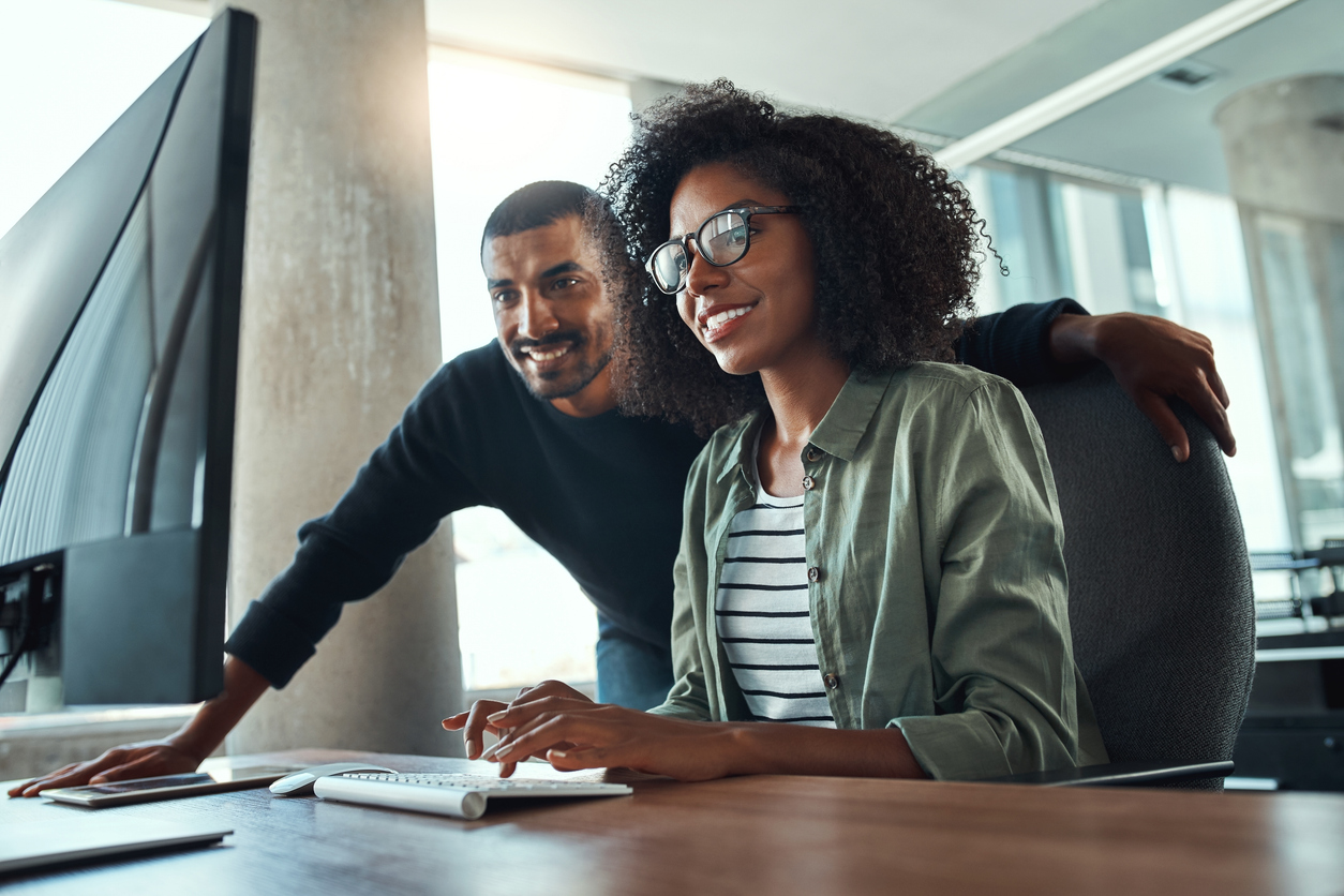 Young businesswoman sitting at her desk working on computer with male colleague standing by